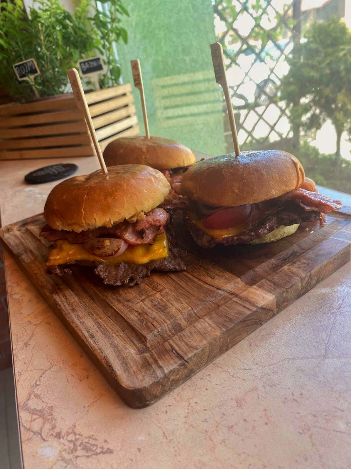 Trzy apetyczne mini burgery z bekonem i serem cheddar, podane na drewnianej desce. W tle zioła w doniczkach i naturalne światło.