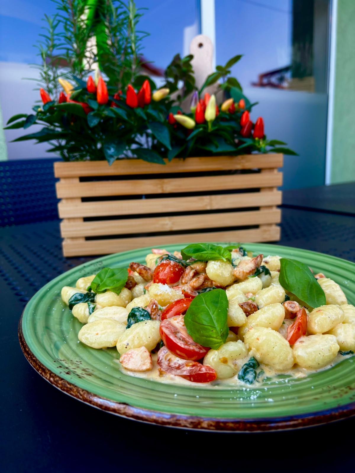 Gnocchi ze szpinakiem i pomidorami na zielonym talerzu, z bazylią. W tle drewniana skrzynka z papryczkami chili i ziołami. Jasne, naturalne światło.