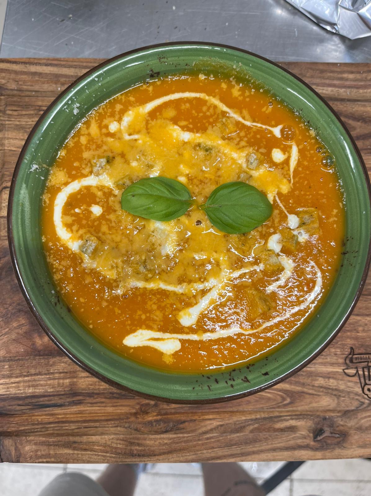 Apetyczna zupa krem pomidorowa w zielonym talerzu, udekorowana kleksem śmietany i listkami bazylii, podana na drewnianej desce. Idealna propozycja na catering.