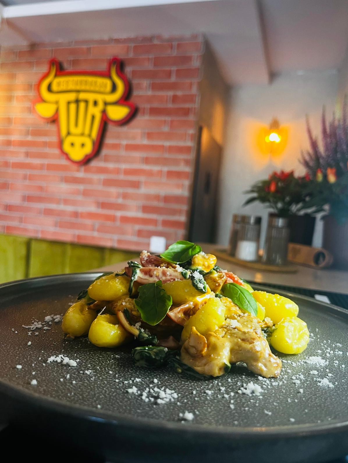 Gnocchi z bazylią na ciemnym talerzu, posypane serem. W tle ceglana ściana z logo restauracji i dekoracje roślinne.
