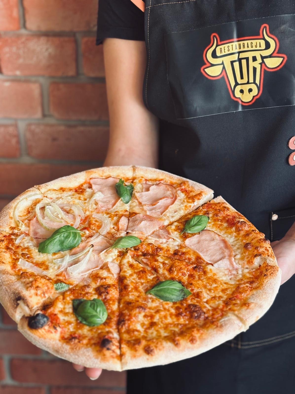 Pizza z szynką, cebulą i bazylią, serwowana na tle ceglanej ściany przez kelnera w Turznie, z logo restauracji na fartuchu. Apetyczny kadr.