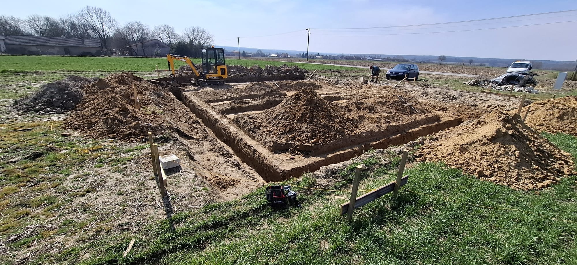 Żółta mini koparka przy wykopie pod fundamenty domu. Widoczny zarys wykopu, usypane pryzmy ziemi, drewniane szalunki i pracownik w tle. Słoneczny dzień.