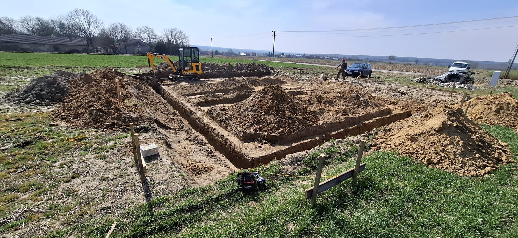 Wykop pod fundamenty z użyciem minikoparki JCB, widoczne wytyczone granice, urobiona ziemia i osoba nadzorująca prace. Prace ziemne w terenie otwartym.