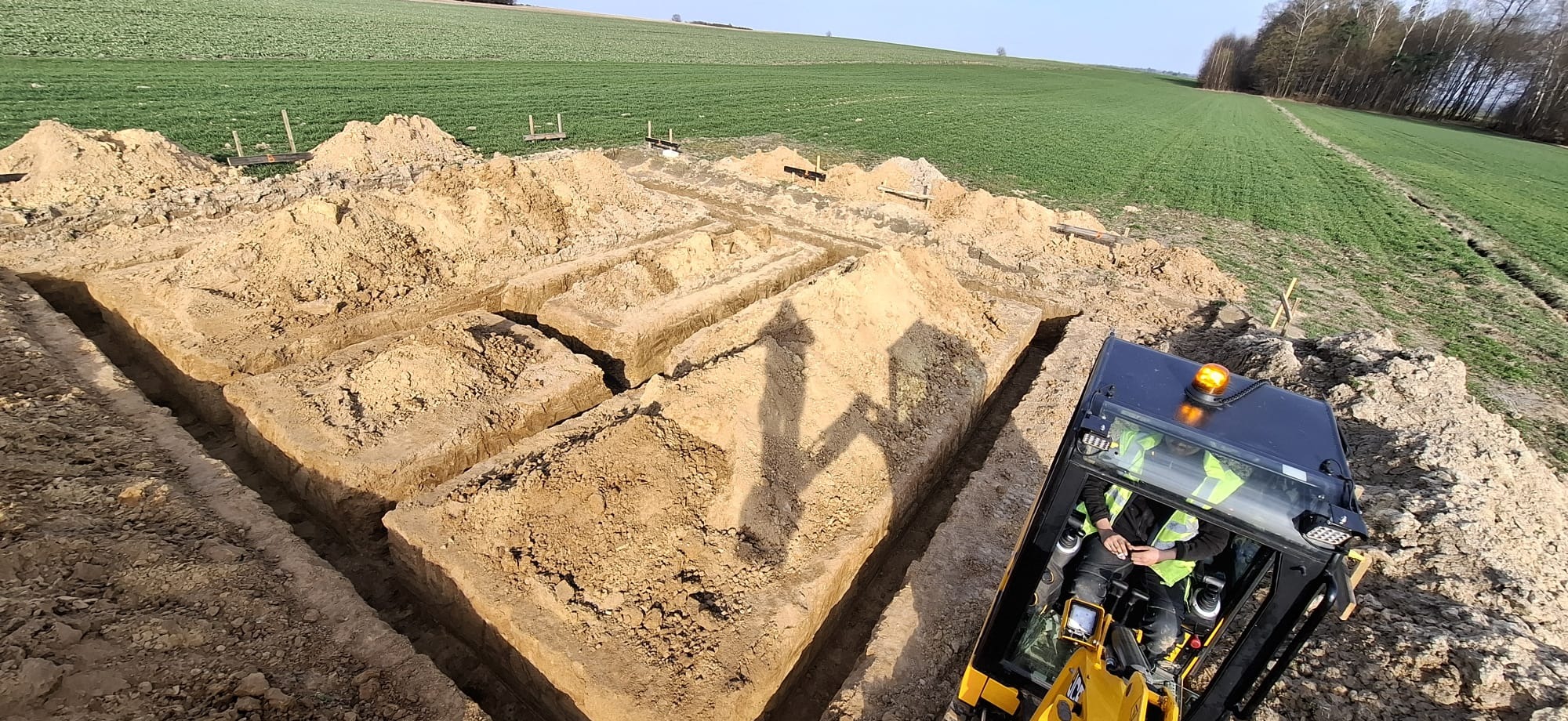 Wykopane rowy pod fundamenty na polu z widoczną mini koparką i operatorem w kabinie. W tle zielone pole i drzewa, słoneczny dzień.