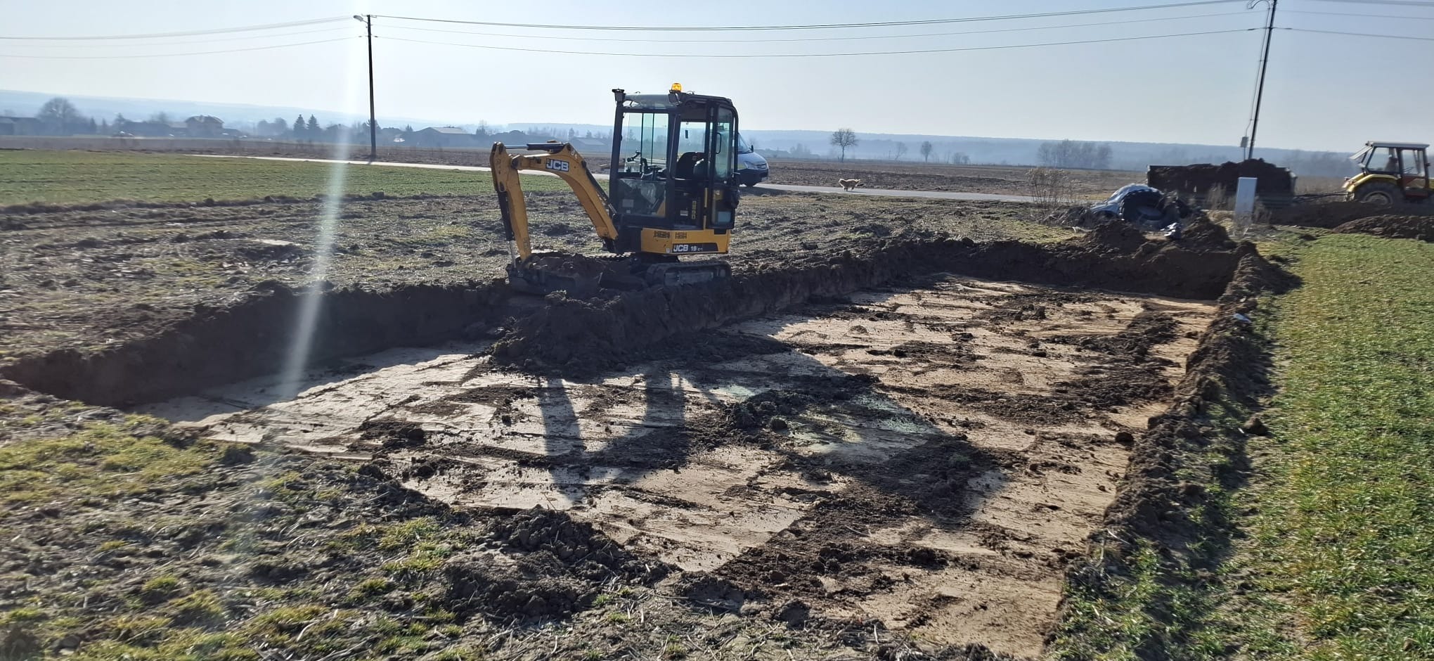 Żółta mini koparka JCB na placu budowy, wykonująca wykop pod fundamenty. Widoczne świeże ślady pracy i wykopany grunt. W tle krajobraz wiejski i błękitne niebo.