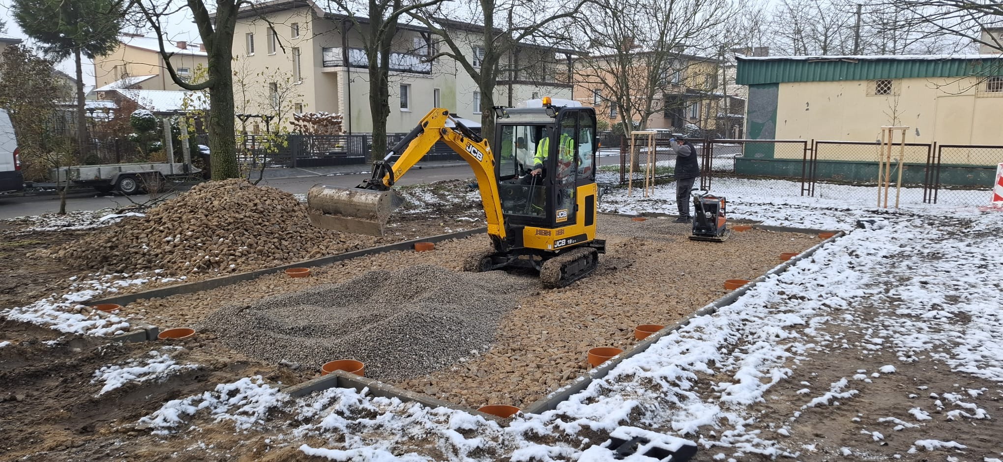 Mini koparka JCB z operatorem wyrównuje teren żwirowy, przygotowując podłoże. W tle widać pryzmę kamieni i zagęszczarkę. Ślady śniegu na ziemi.