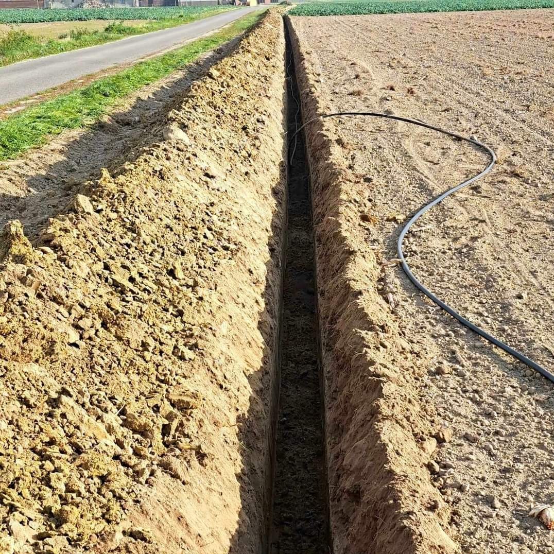 Wąski wykop w ziemi, z widocznym czarnym kablem wewnątrz. Po lewej droga, po prawej pole uprawne. Wykop świeżo wykonany, ziemia jasnobrązowa.