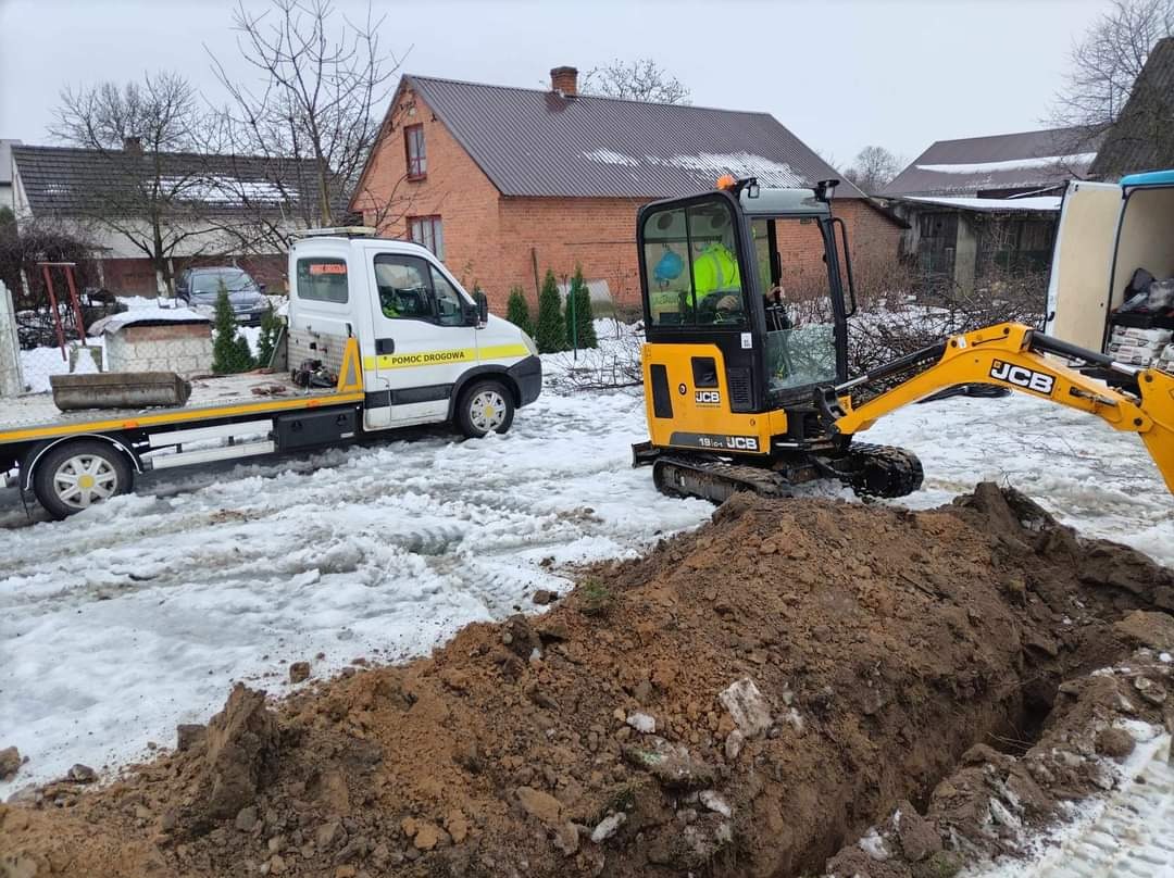 Żółta mini koparka JCB 19C-1 w trakcie prac ziemnych, widoczny wykop w zasypanym śniegiem terenie. Obok laweta pomocy drogowej.