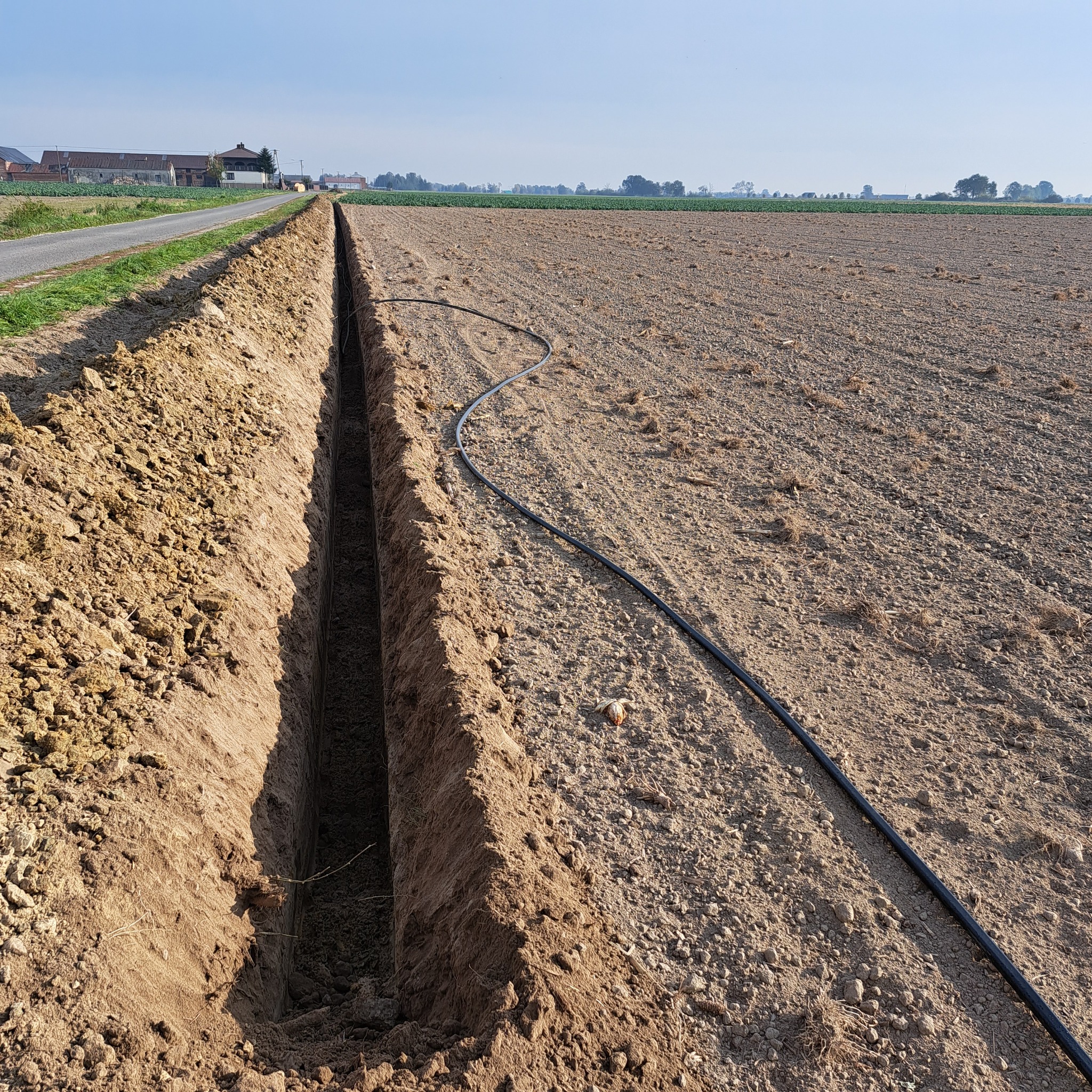 Długi, wąski wykop w polu z czarną rurą na powierzchni, prawdopodobnie pod instalacje. Widok na odległe zabudowania i niebo.