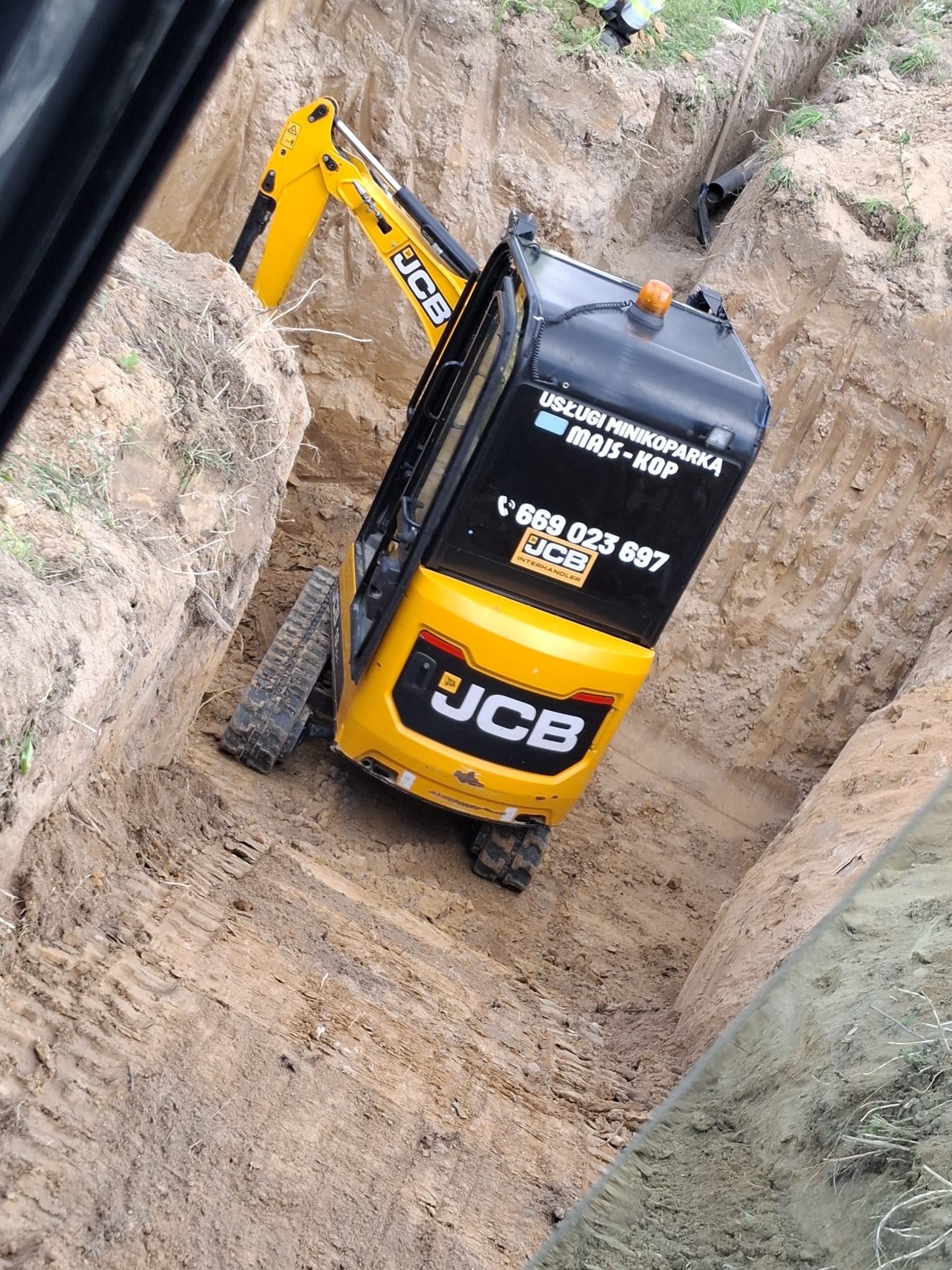 Żółta mini koparka JCB w wykopie, widoczne rury i operator. Wykop wąski, prawdopodobnie pod instalacje. Na koparce naklejka z numerem telefonu.