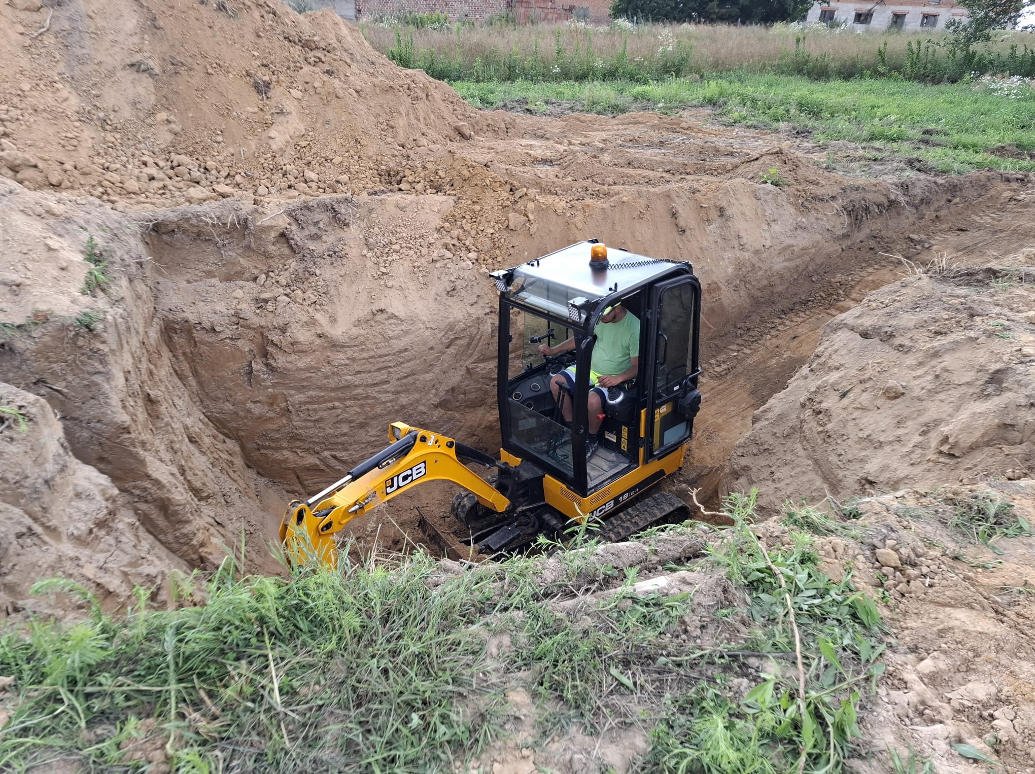 Żółta mini koparka JCB w głębokim wykopie na działce, operator w kabinie. W tle hałda ziemi i budynek. Prace ziemne w toku.