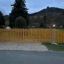 Żółte ogrodzenie z pionowych desek i metalowych słupków na tle jesiennego krajobrazu z górami i domem. U podstawy ogrodzenia wysypany żwir.