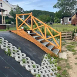 Drewniane schody z balustradą prowadzące na podwyższony teren, z metalowymi stopniami. Na zboczu betonowe ażurowe płyty z roślinnością, w tle dom i zieleń.