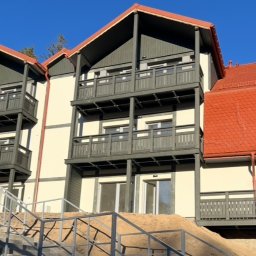 DrewMax - Trzykondygnacyjny budynek z drewnianymi balustradami balkonowymi w kolorze grafitowym i schodami wejściowymi z metalową barierką. Czerwony dach i jasna elewacja.