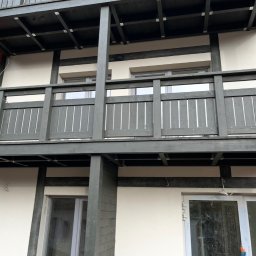 DrewMax - Dwupoziomowy balkon z drewnianą balustradą w odcieniach szarości, widok z dołu, z fragmentem dachu i elewacją budynku w tle. Detal konstrukcji.