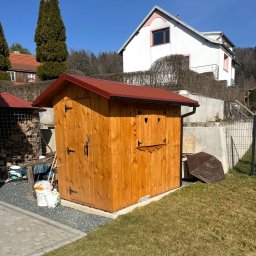DrewMax - Drewniana budka narzędziowa z czerwonym dachem, ozdobiona wyciętymi sercami, na tle domu i zieleni. Widoczne detale wykończenia i otoczenie.