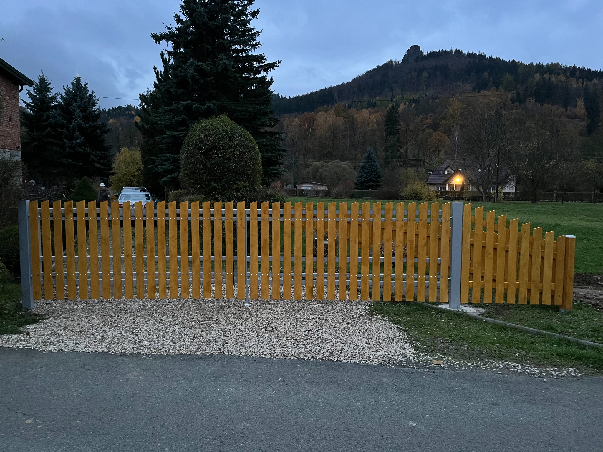 Żółte ogrodzenie z pionowych desek i metalowych słupków na tle jesiennego krajobrazu z górami i domem. U podstawy ogrodzenia wysypany żwir.