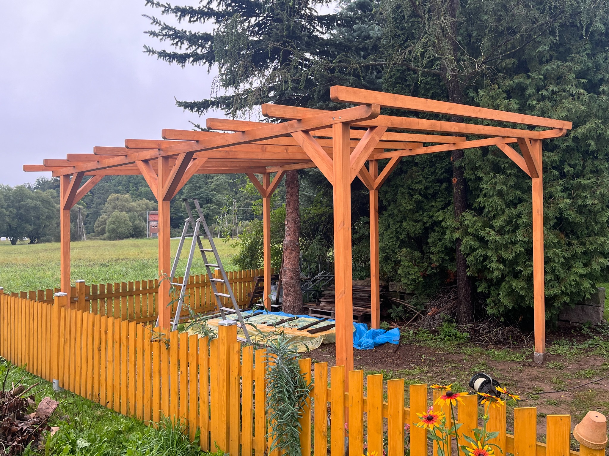 Montaż drewnianej pergoli w ogrodzie, widoczna drabina, narzędzia i elementy konstrukcyjne. Żółte ogrodzenie i zieleń w tle. Etap budowy z bliska.