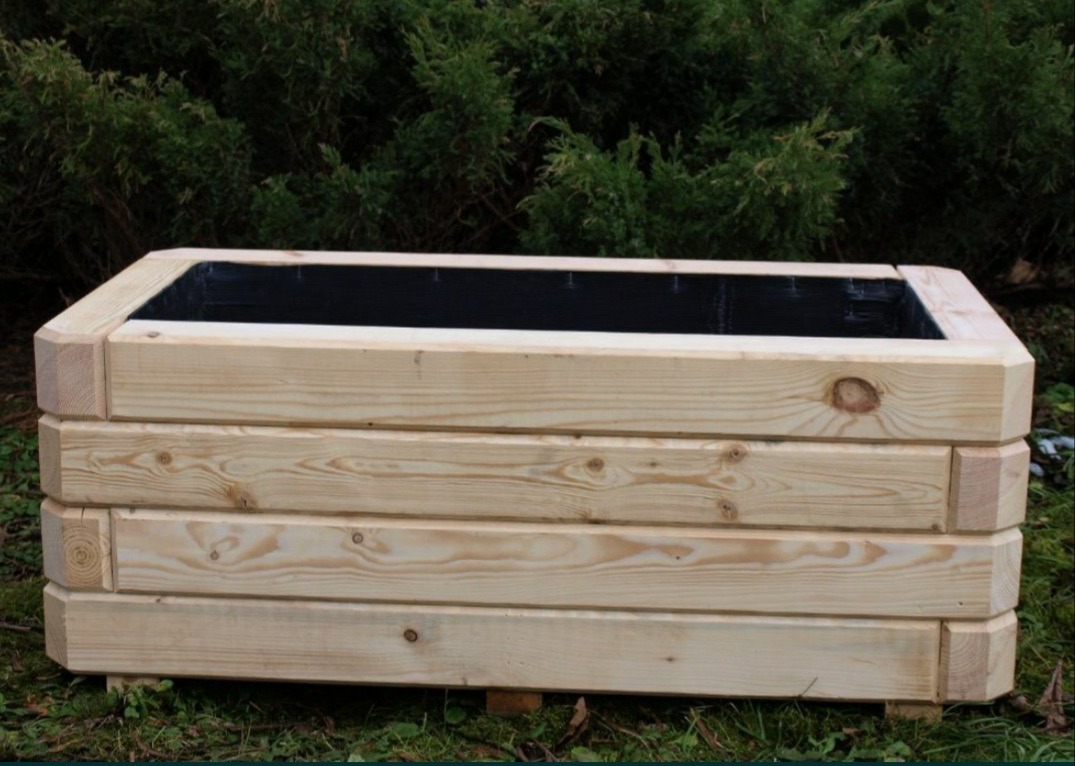 Prostokątna, drewniana skrzynia na rośliny z czarnym wnętrzem, wykonana z jasnych desek, na tle zielonych krzewów. Solidna konstrukcja, naturalne drewno.
