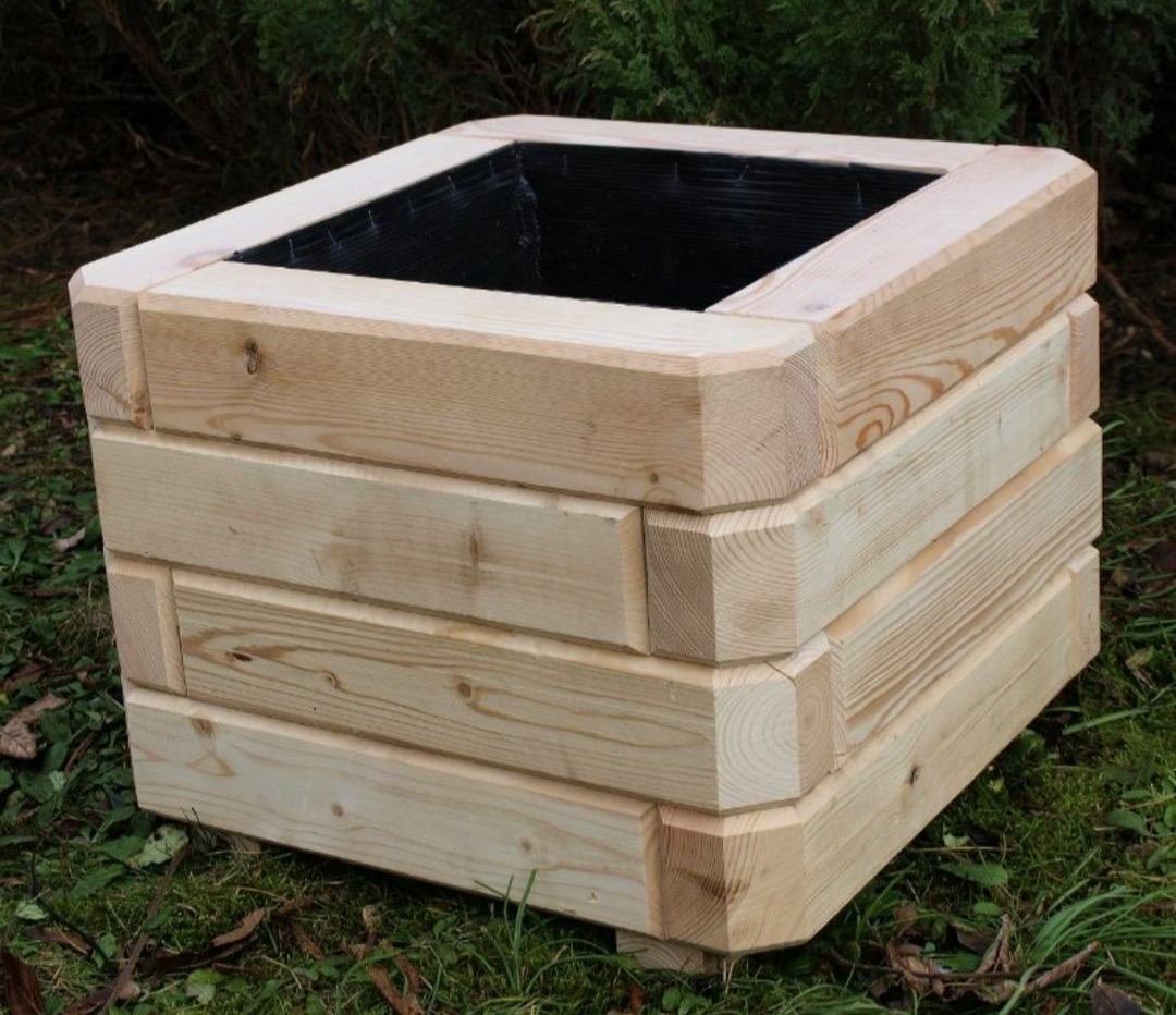 Kwadratowa, drewniana donica z jasnego drewna, złożona z czterech warstw desek, stoi na trawie na tle zieleni. Wnętrze wyłożone czarną folią.