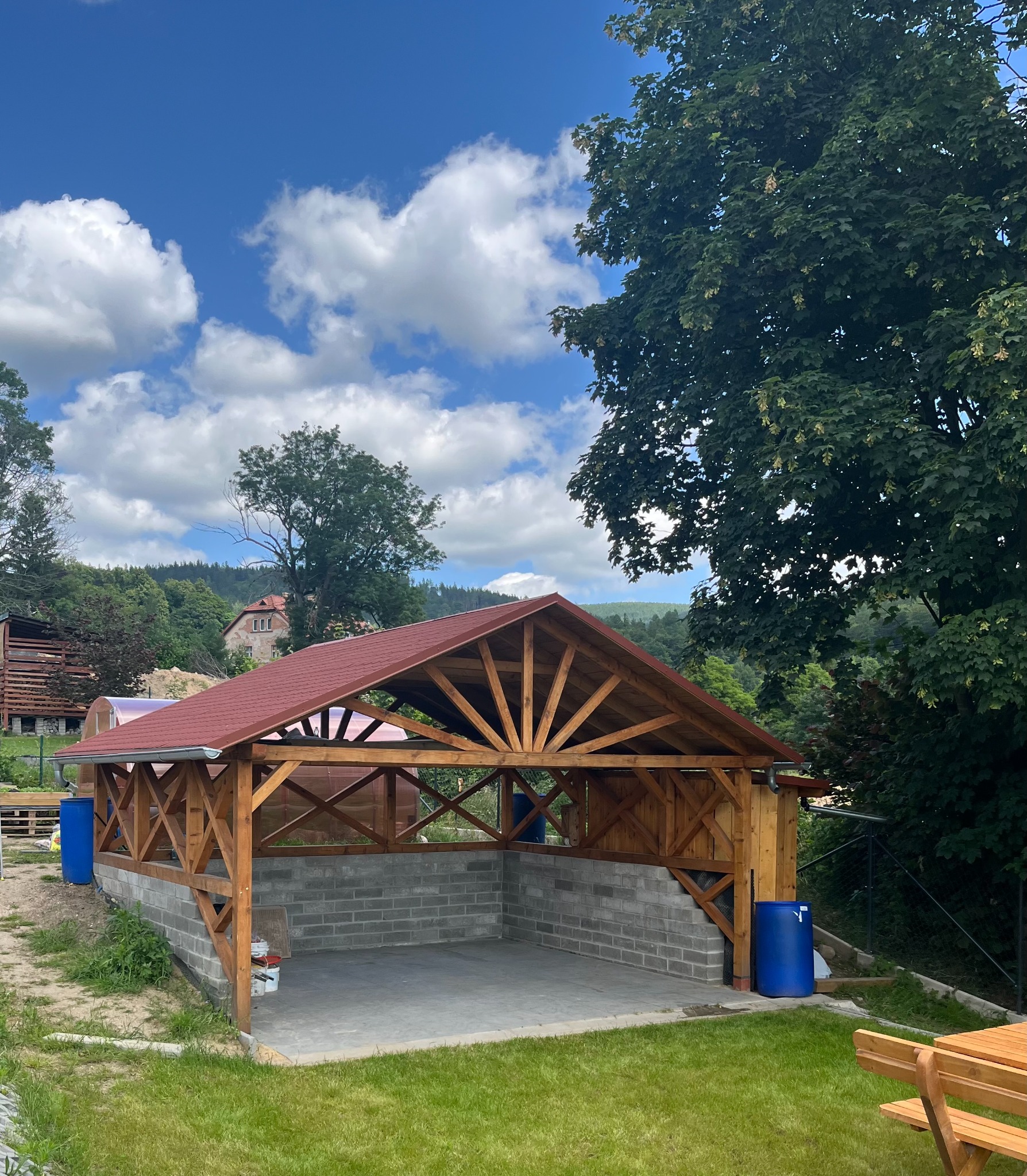 Drewniana altana z czerwoną dachówką na fundamencie z szarych bloczków, otoczona zielenią i drzewami w słoneczny dzień. Widoczne niebieskie beczki.