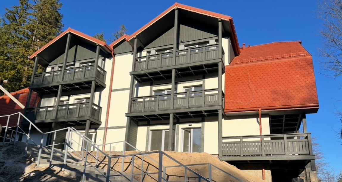 Trzykondygnacyjny budynek z drewnianymi balustradami balkonowymi w kolorze grafitowym i schodami wejściowymi z metalową barierką. Czerwony dach i jasna elewacja.