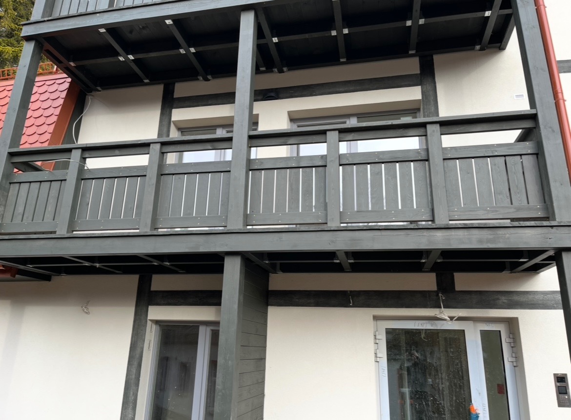 Dwupoziomowy balkon z drewnianą balustradą w odcieniach szarości, widok z dołu, z fragmentem dachu i elewacją budynku w tle. Detal konstrukcji.