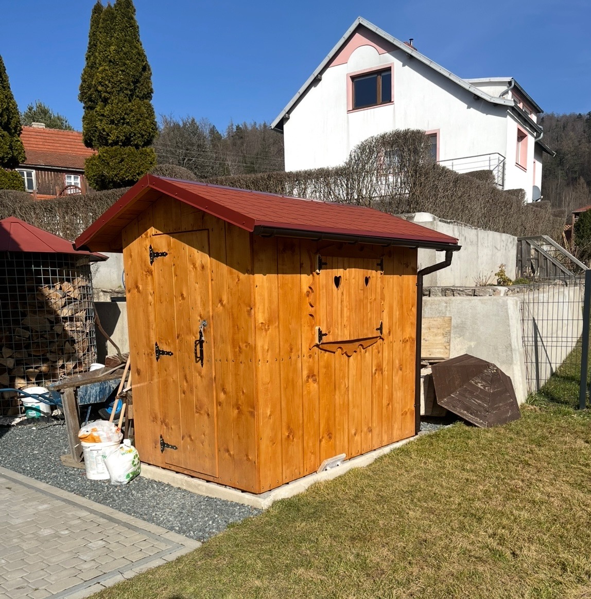 Drewniana budka narzędziowa z czerwonym dachem, ozdobiona wyciętymi sercami, na tle domu i zieleni. Widoczne detale wykończenia i otoczenie.