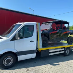 Biała laweta z czarno-czerwonym pojazdem Polaris General 1000 na platformie, zaparkowana na brukowanej powierzchni, w tle czerwony budynek i błękitne niebo.