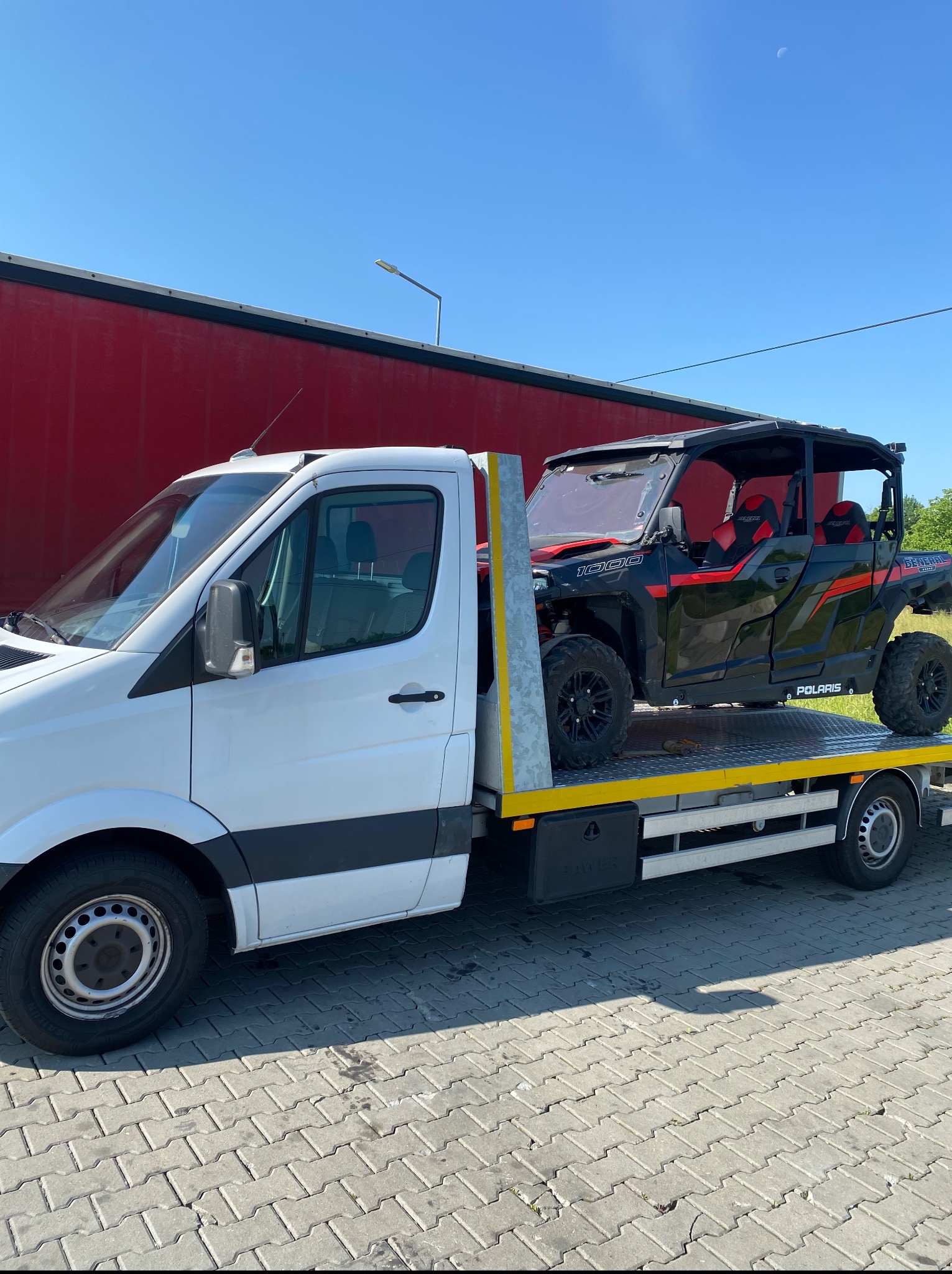 Biała laweta z czarno-czerwonym pojazdem Polaris General 1000 na platformie, zaparkowana na brukowanej powierzchni, w tle czerwony budynek i błękitne niebo.