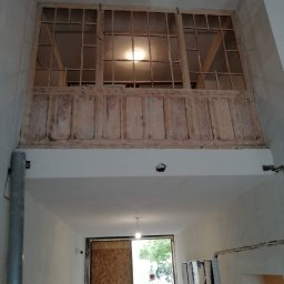 PacBour-Styl Nataliia Kostrova - Wnętrze z drewnianą konstrukcją antresoli z oknami i panelami, widok z dołu na otwarte drzwi i postać. Widać surowe ściany i instalacje.