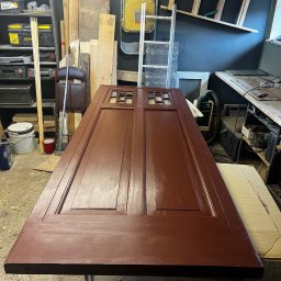 PacBour-Styl Nataliia Kostrova - Odnowione, brązowe drzwi z szybkami leżące na stole w warsztacie, w tle drabiny i narzędzia. Widoczny świeży lakier na powierzchni drzwi.