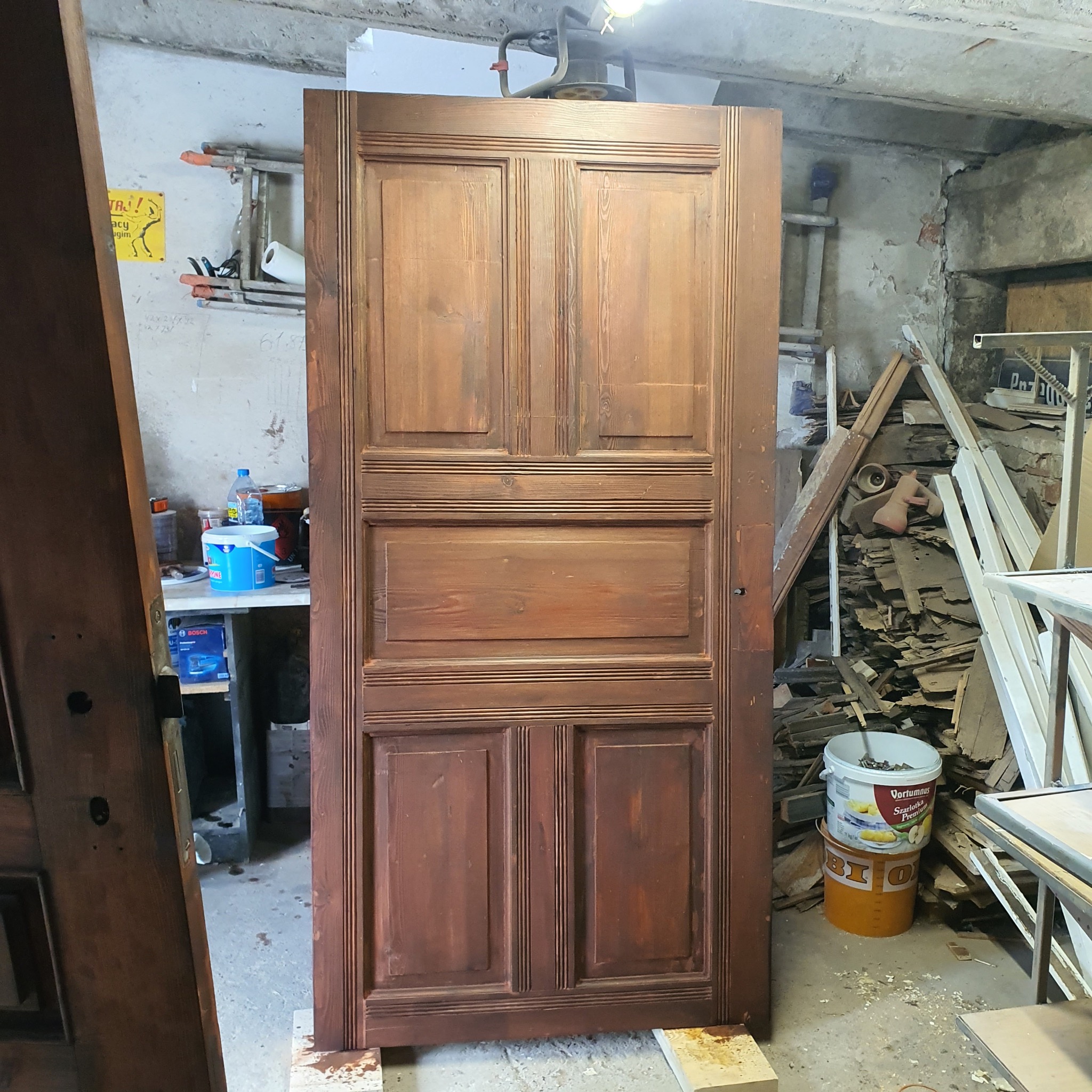 Odrestaurowane, drewniane drzwi z płycinami, ustawione pionowo w warsztacie. Widoczne ślady renowacji i charakterystyczne detale stolarskie. Oświetlenie górne.