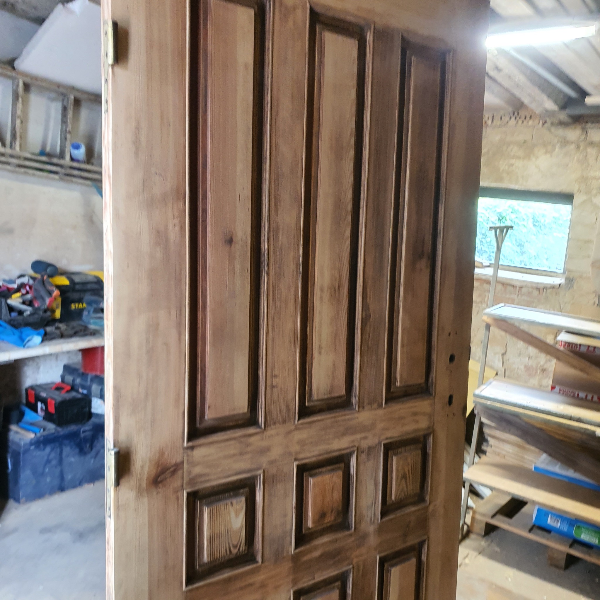 Odnowione drewniane drzwi w warsztacie. Widoczne detale rzeźbienia i bejcowania. W tle narzędzia i materiały stolarskie, okno z widokiem na zieleń.