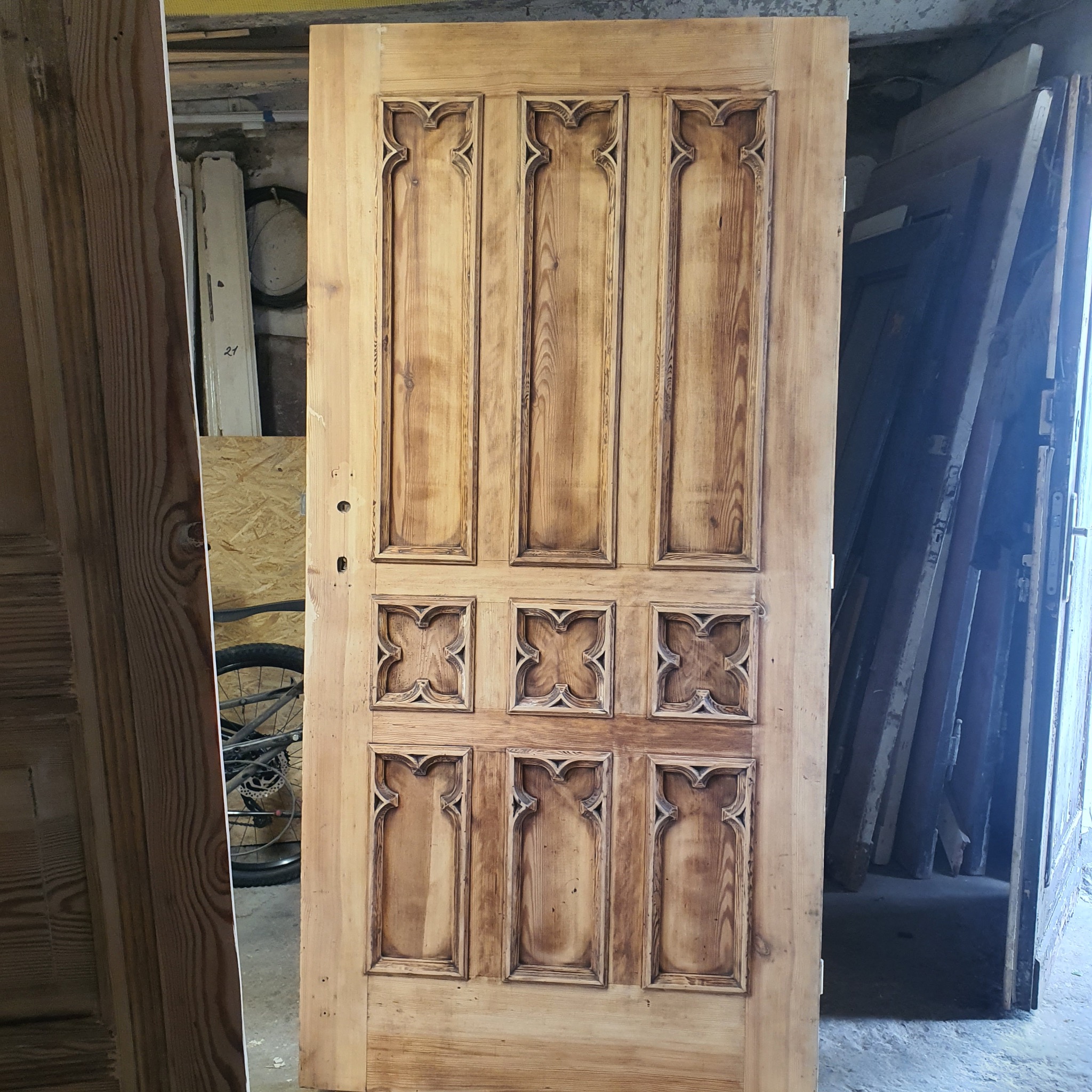 Odrestaurowane, drewniane drzwi z dekoracyjnymi, gotyckimi panelami, ustawione pionowo w warsztacie. Widoczne ślady renowacji i surowe drewno. W tle inne elementy stolarki.