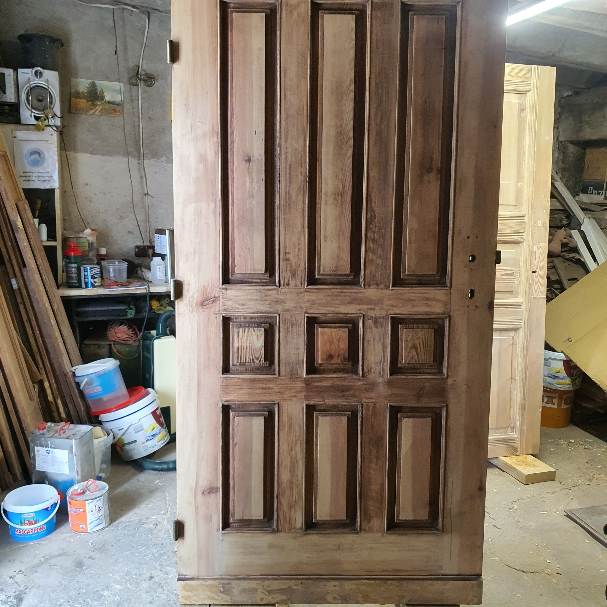 Drewniane drzwi po renowacji z widocznymi detalami rzeźbienia i bejcowania, stojące w warsztacie. W tle farby, deski i inne drzwi w trakcie odnawiania.
