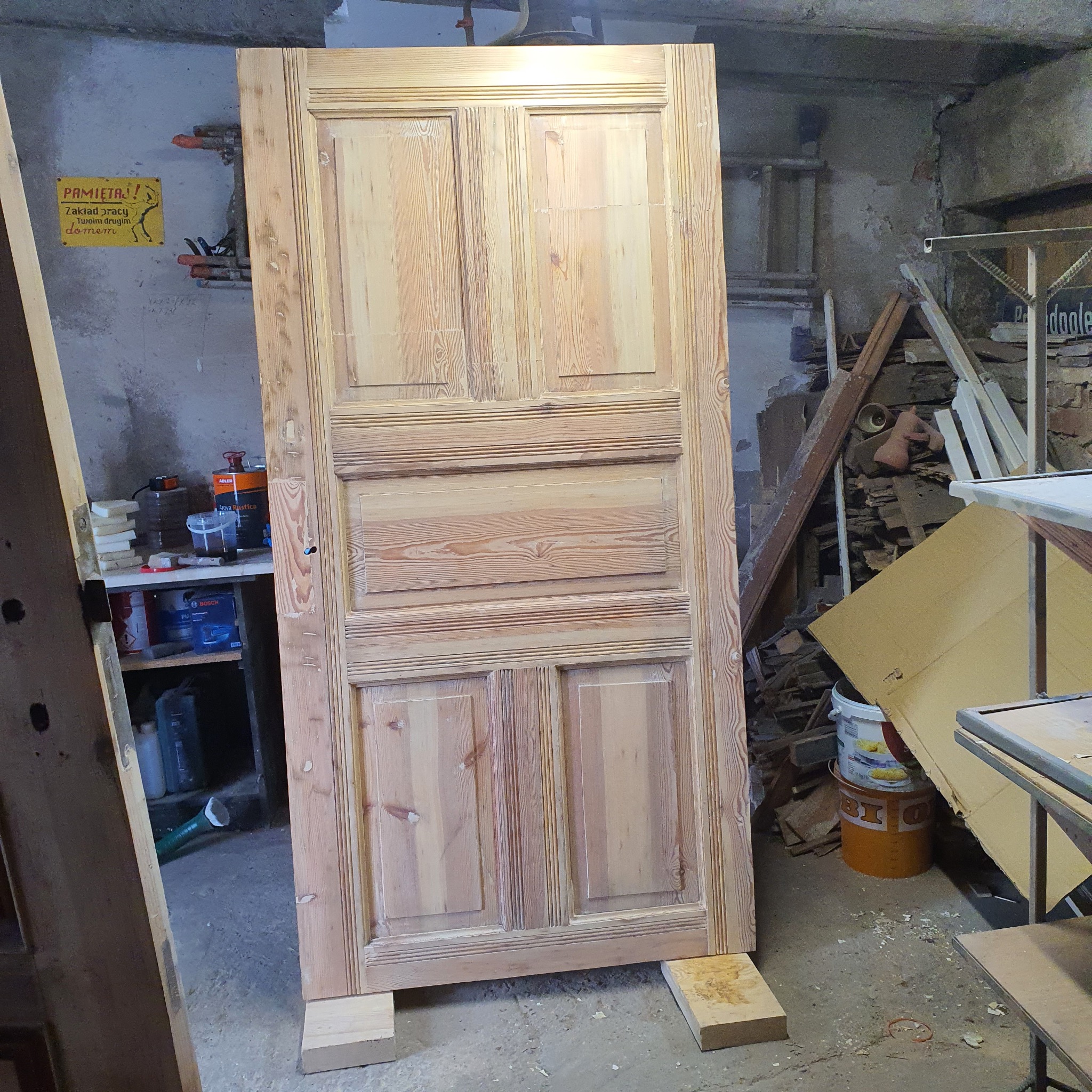 Drewniane, odrestaurowane drzwi wewnętrzne z czterema płycinami, ustawione w warsztacie na drewnianych klockach. Widoczne narzędzia i materiały stolarskie w tle.