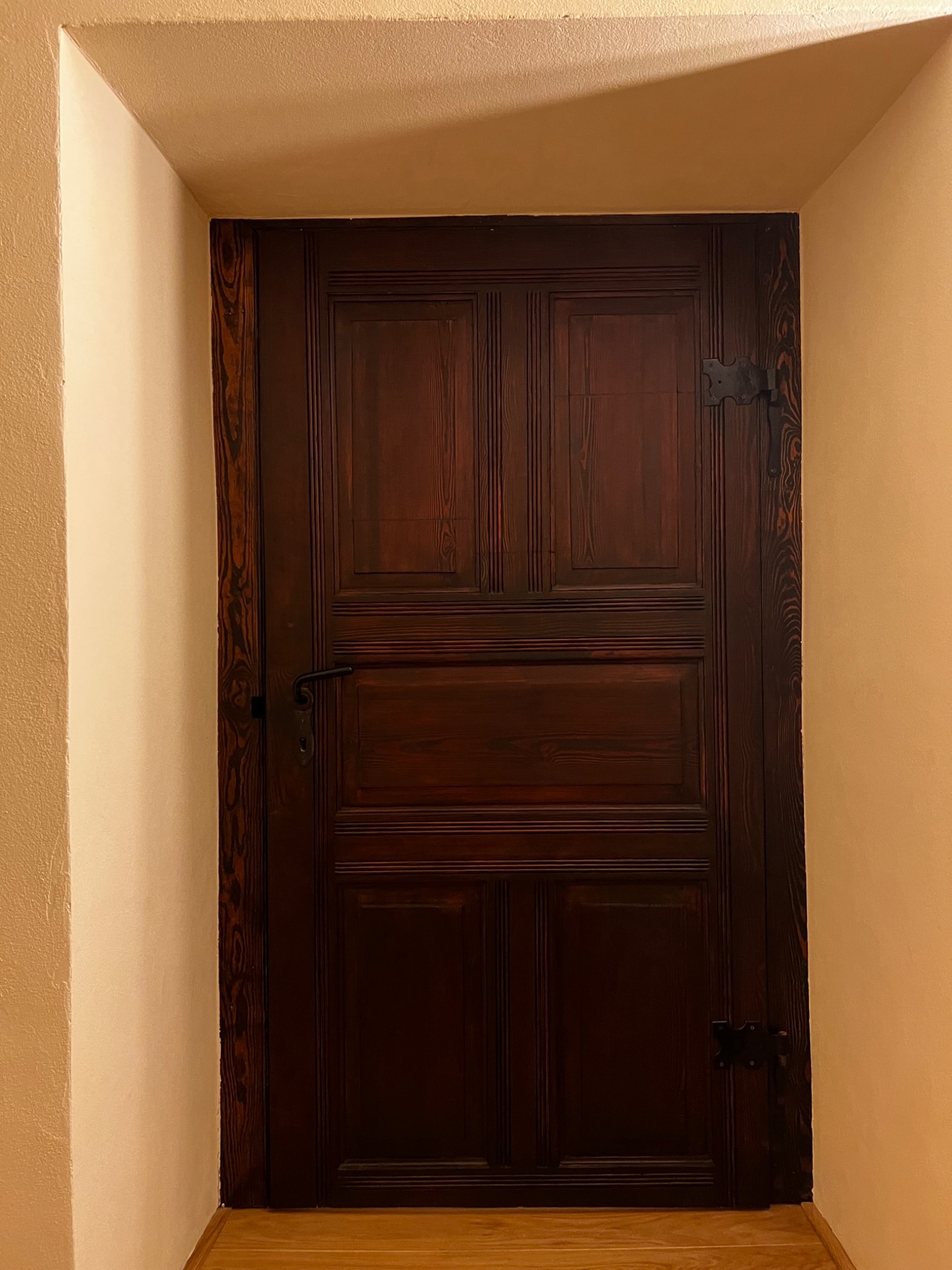 Ciemne, drewniane drzwi z metalowymi zawiasami w beżowej ścianie. Widoczny fragment drewnianej podłogi. Klasyczny styl wykończenia wnętrza, ciepłe oświetlenie.