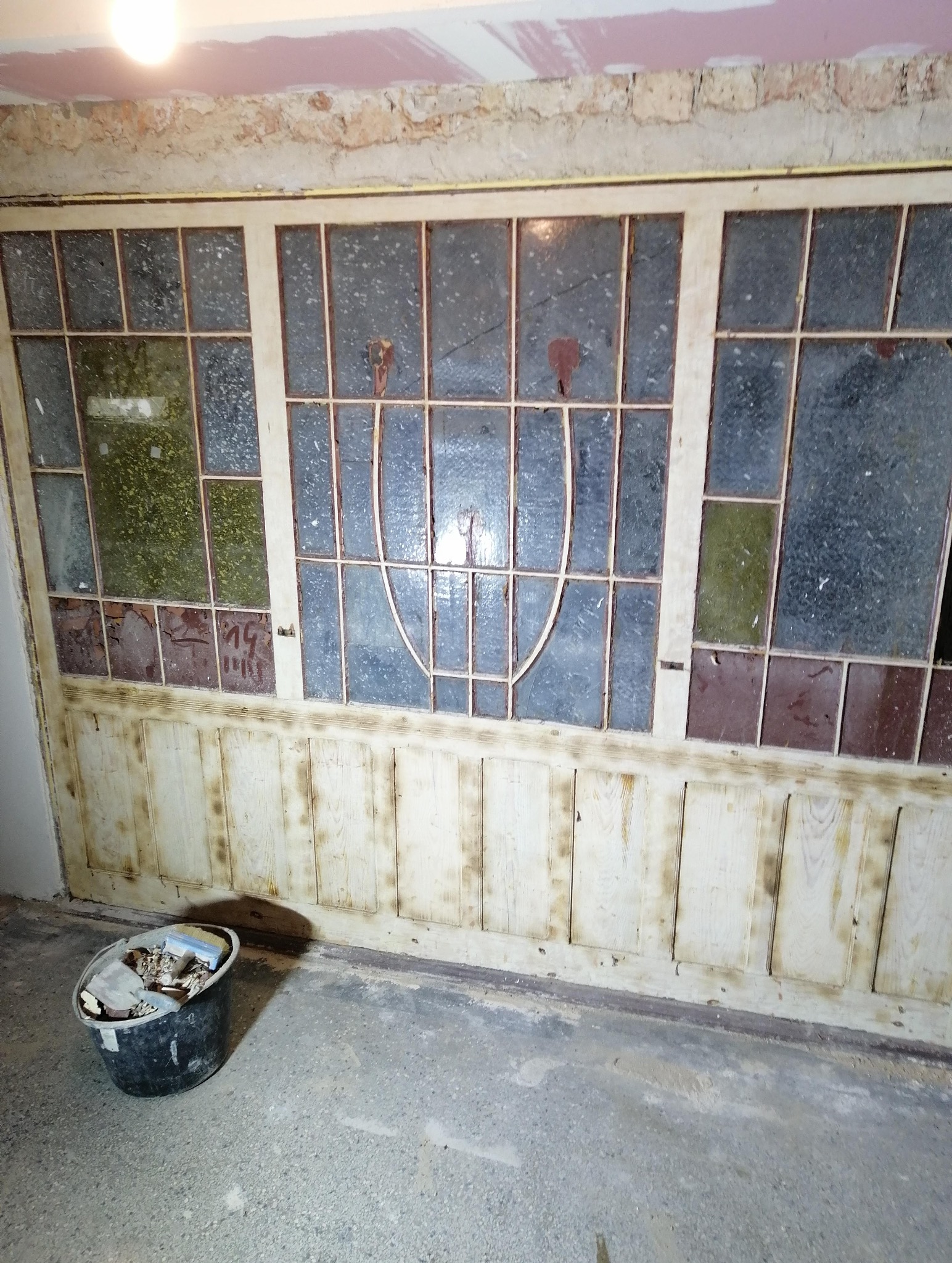 Stara, drewniana ściana z szybami w trakcie renowacji. Widoczne ślady farby, szpachli i wiadro z odpadami. Górna część ściany odsłonięta, przygotowana do dalszych prac.