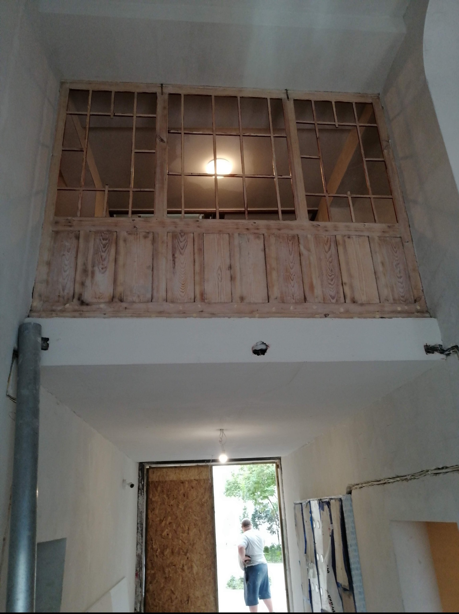 Wnętrze z drewnianą konstrukcją antresoli z oknami i panelami, widok z dołu na otwarte drzwi i postać. Widać surowe ściany i instalacje.