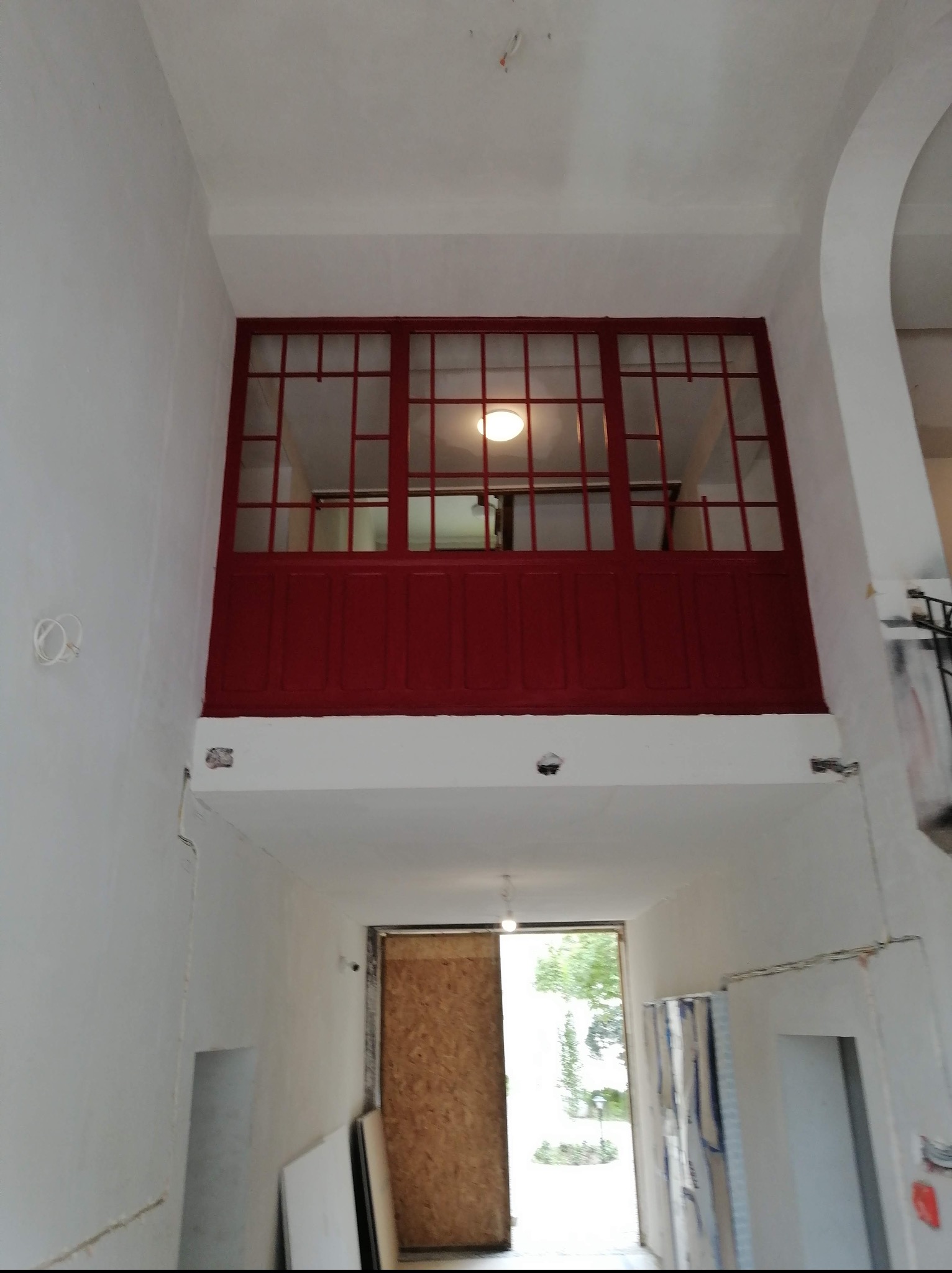 Wnętrze w trakcie remontu z widoczną antresolą z czerwoną balustradą. Ściany białe, widoczne ślady po instalacjach. W tle zamknięte drzwi i widok na zieleń.