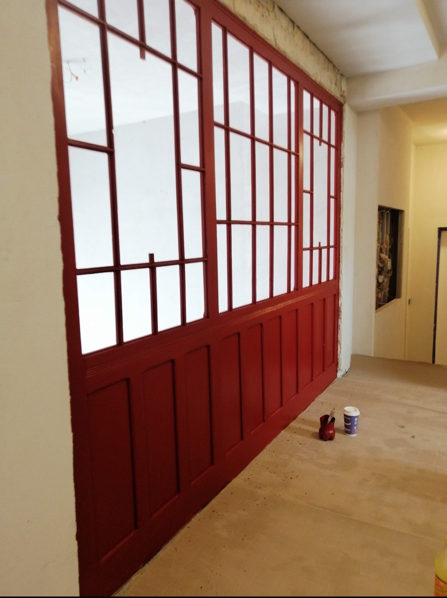 Czerwona, przeszklona ścianka działowa z drewnianą ramą, widoczna z boku. Na podłodze akcesoria malarskie. W tle jasne pomieszczenie.