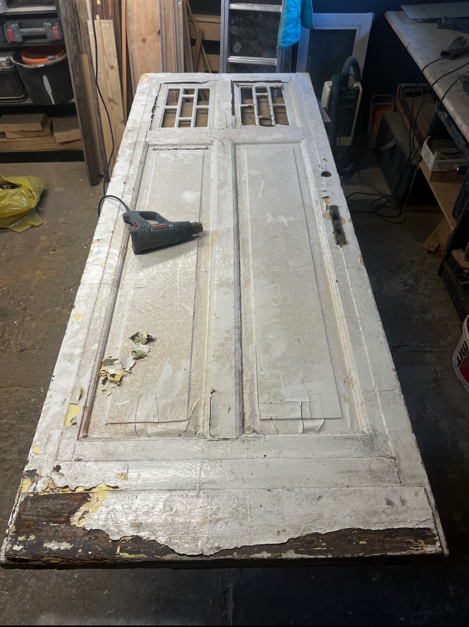 Stara, drewniana, biała skrzydło drzwiowe z odchodzącą farbą leży na stole w warsztacie. Obok opalarka i resztki farby. Renowacja w toku.