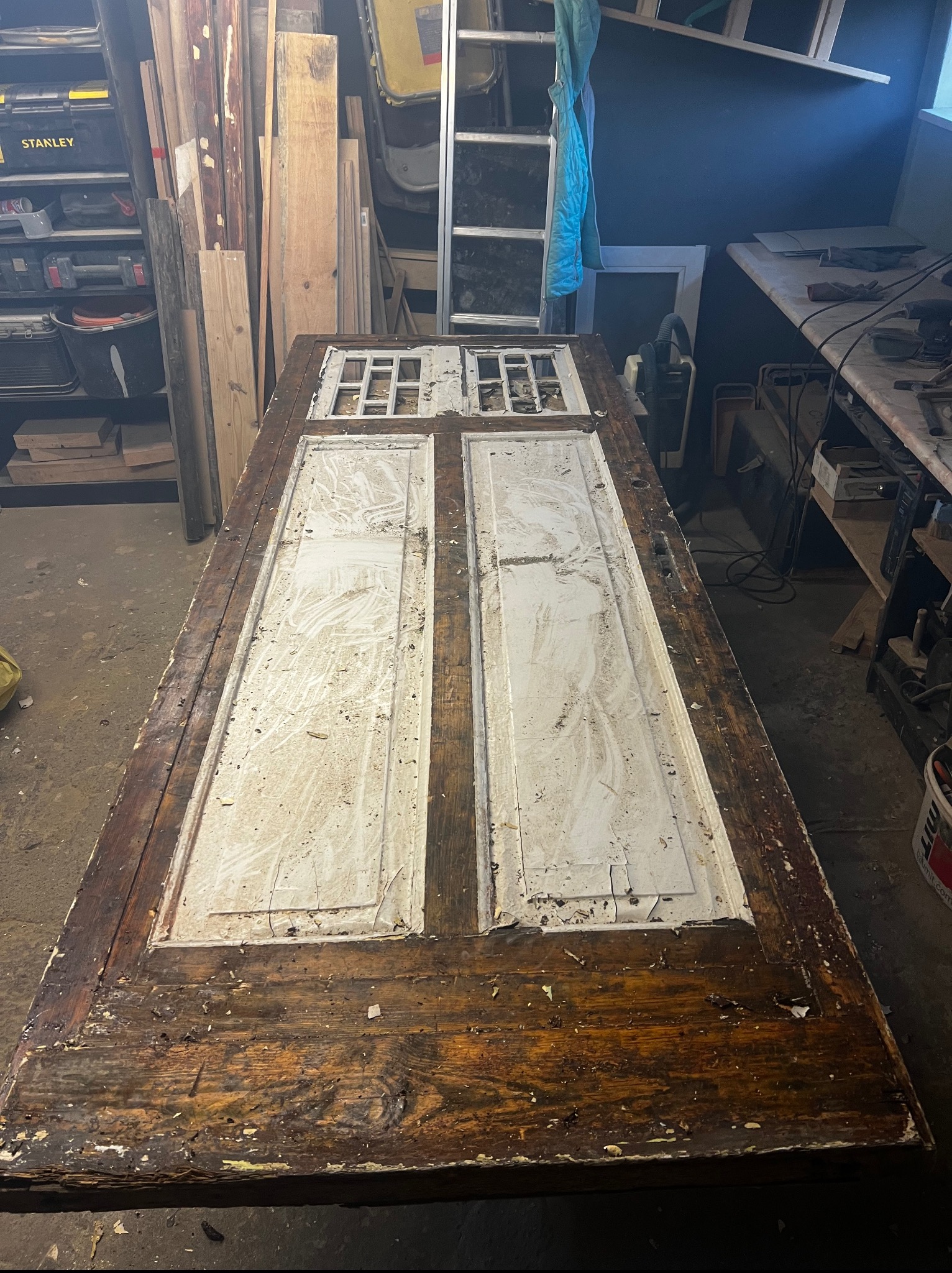 Stara, drewniana rama drzwiowa z resztkami białej farby leżąca na stole w warsztacie, gotowa do renowacji. Widoczne ślady użytkowania i odpryski farby.