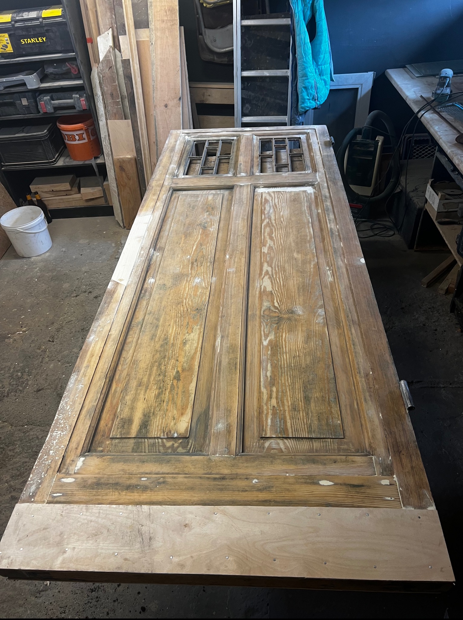 Stara, drewniana, dwuskrzydłowa brama z kratką w górnej części, leżąca na stole w warsztacie. Widać ślady farby i renowacji, oraz nową listwę u dołu.