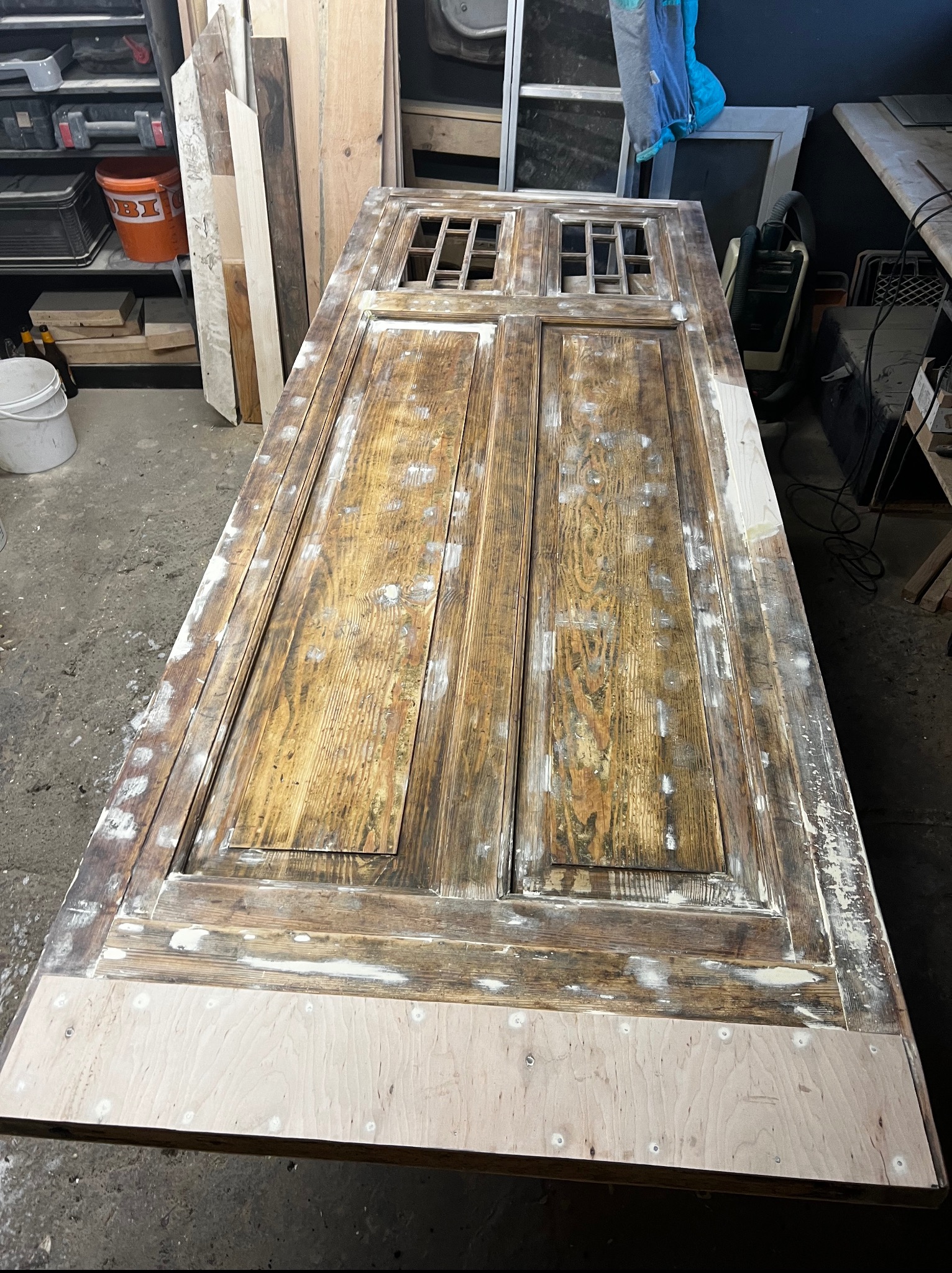 Stara, drewniana, dwuskrzydłowa brama z okienkami po renowacji, leżąca na stole. Widoczne ślady szpachli i surowe drewno u dołu. W tle warsztat.