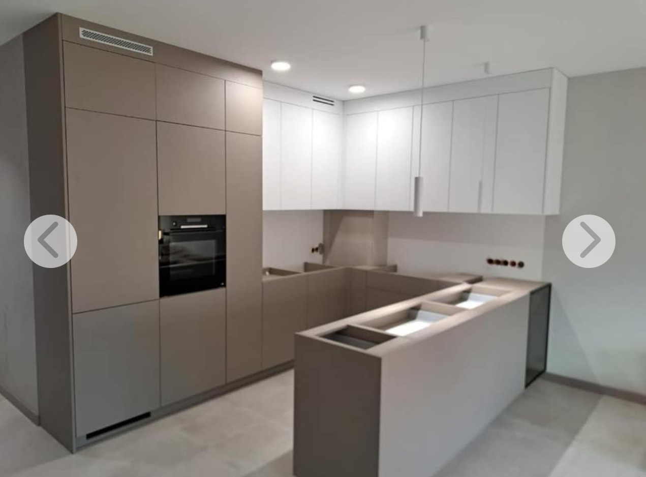 Nowoczesna kuchnia w odcieniach szarości i bieli z minimalistycznym oświetleniem. Częściowo zamontowana wyspa kuchenna z miejscem na zlew. Widoczny piekarnik.