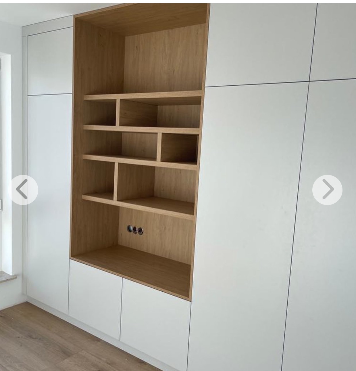 Nowoczesna szafa na wymiar z białymi frontami i drewnianym regałem z półkami. Widoczne gniazdka elektryczne wewnątrz regału. Minimalistyczny design, jasne wnętrze.