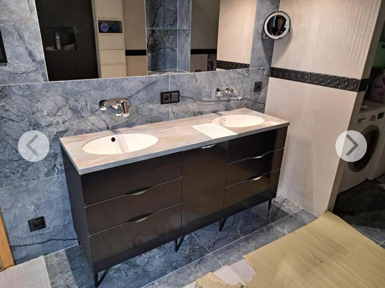 Nowoczesna łazienka z podwójną umywalką na szafce z ciemnego drewna, marmurowy blat, szare płytki imitujące kamień i okrągłe lustro z podświetleniem.