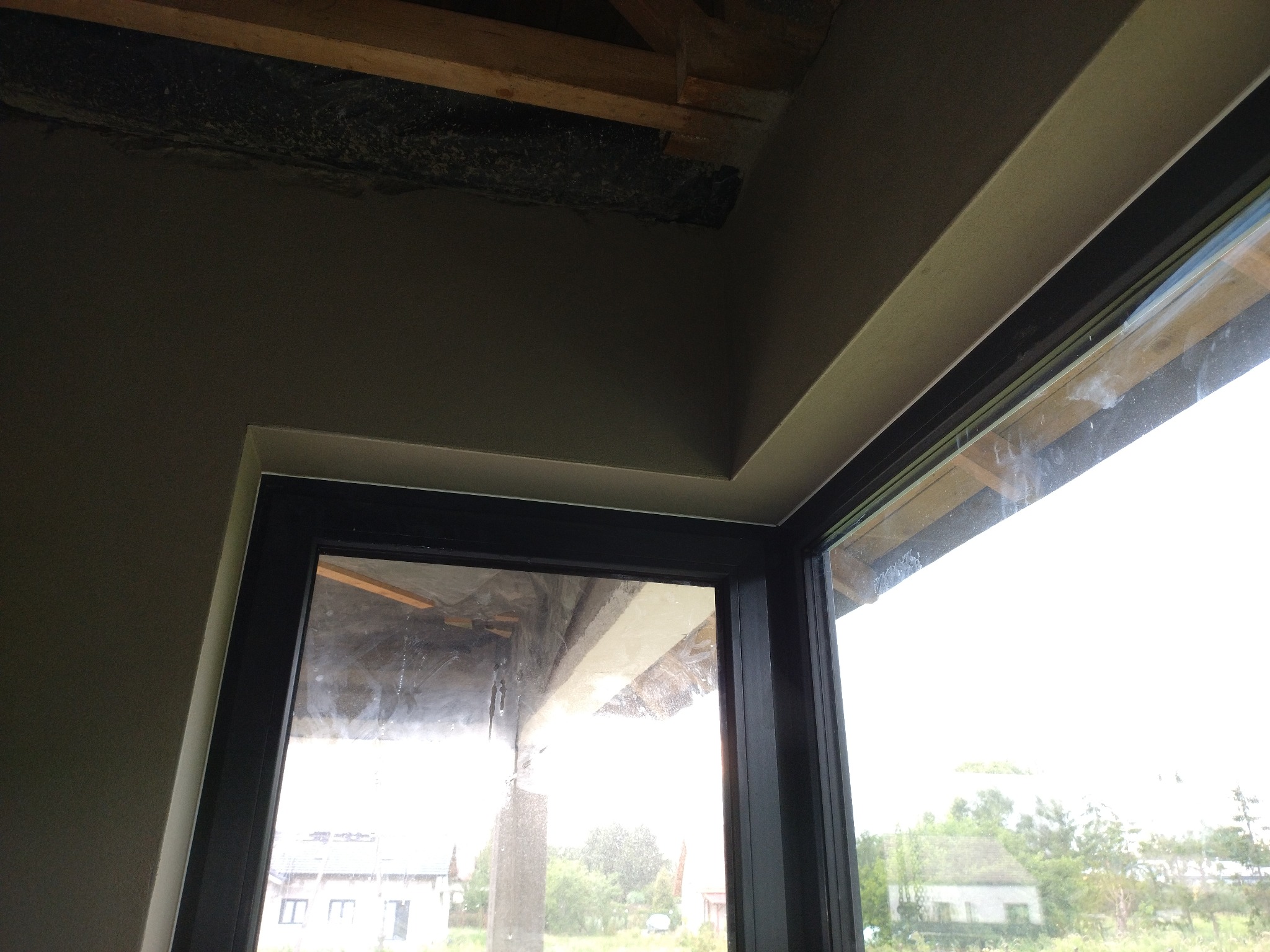 Wnętrze po tynkowaniu: gładkie, szare ściany z czarną ramą okienną i widokiem na zewnątrz. Detal wykończenia sufitu i okna.